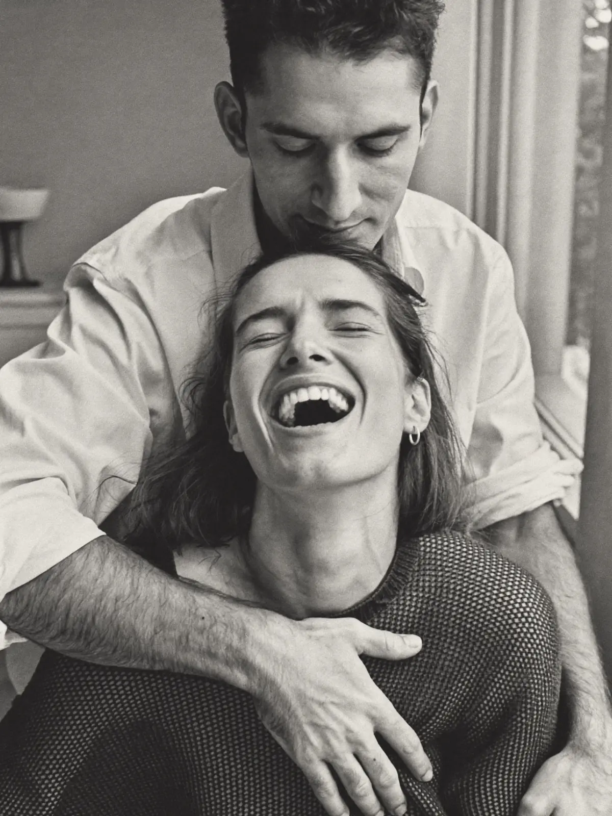 A man in a white shirt hugs a laughing woman from behind by a window, black and white