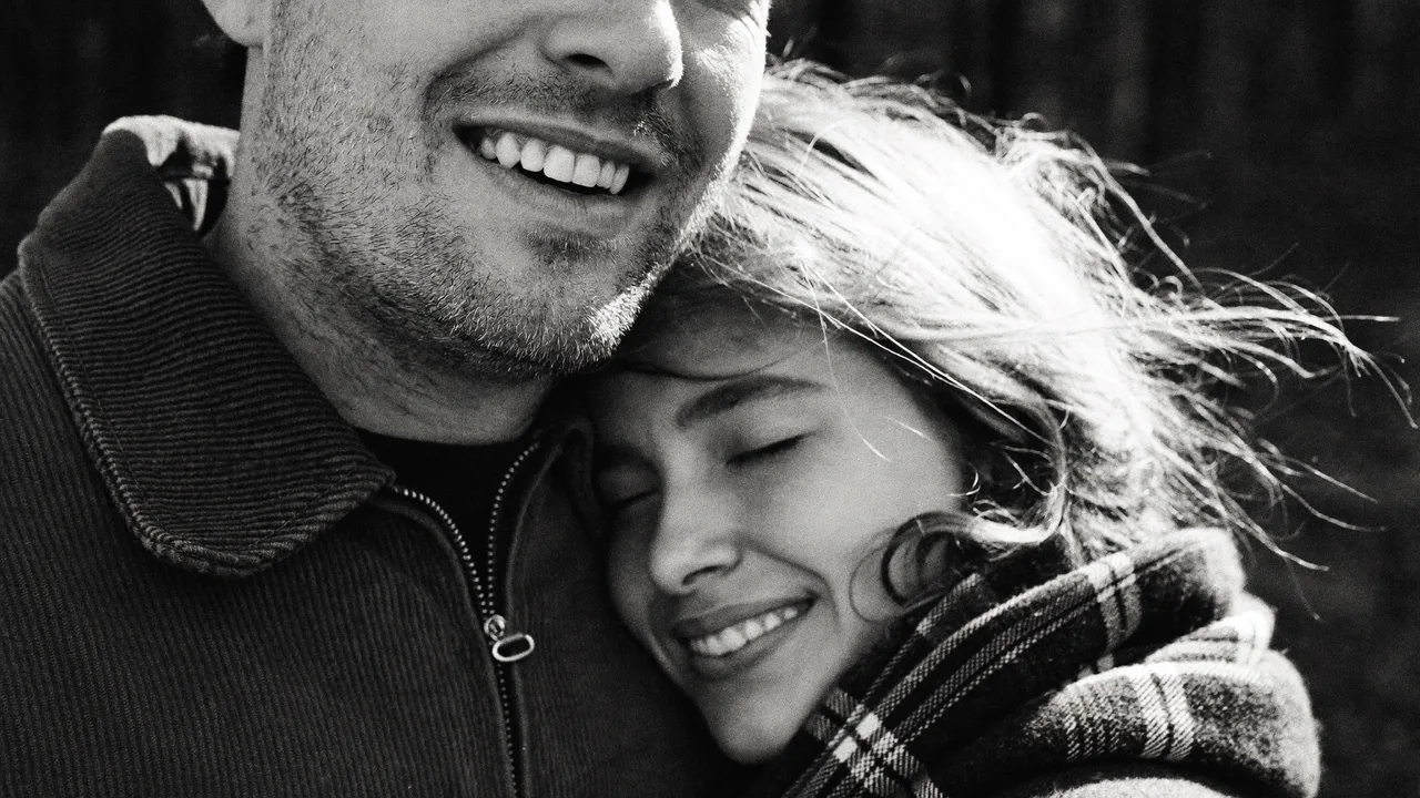 A smiling man holds a woman with windblown hair in a plaid scarf, black and white
